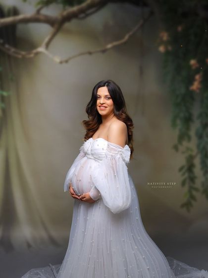 An ethereal portrait of a mother-to-be in a white pearl-studded gown. The dreamy, enchanted forest backdrop adds to the magical quality of the image.