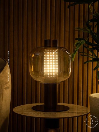 This table lamp features a perforated inner cylinder shielded by an outer amber glass shade. The design creates a mesmerizing pattern of light, offering a warm and intricate glow for a side table or console.