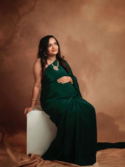 A full-length portrait of a mom-to-be in a beautiful green saree, seated on a simple white block. The classic pose and painterly backdrop create a timeless feel.