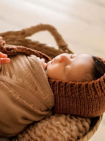 This portrait highlights a cozy knit bonnet and a matching wrap. The textures of the woven basket and soft fabrics add depth and warmth to the image.