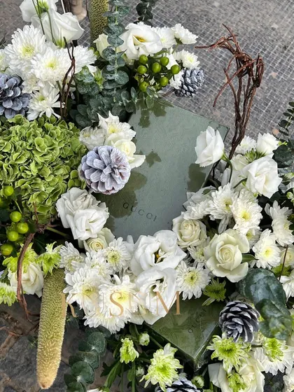 Dressing your gifts with flowers, this time in chic whites and greens. A luxury gift box is nestled within a bed of white roses, green hydrangeas, and rustic pinecones, creating an elegant and cohesive presentation.