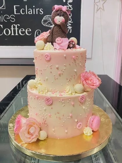 A two-tiered pink and white cake with a cute teddy bear on top. The cake is elegantly decorated with sugar pearls, buttons, macarons, and fondant roses.