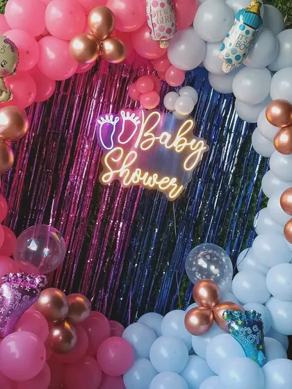 This is a close-up of a popular baby shower decoration. It features a full ring of pink and blue balloons, a shiny backdrop, a "Baby Shower" neon sign, and cute baby-themed foil balloons to complete the look.