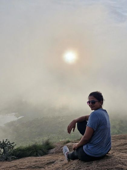 A trekker sits and smiles, with the sun breaking through the clouds at Uttari Betta.