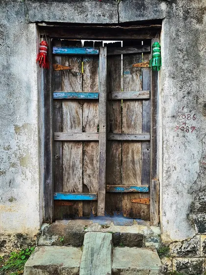 A rustic wooden door held together by brightly colored patches of blue paint. The small, handmade tassels hanging above add a personal touch to this weathered and repaired entryway.