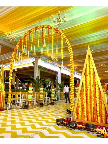 An entrance arch for a Haldi ceremony, decorated with marigold garlands and traditional floral arrangements. The yellow and white chevron flooring adds a modern touch to the traditional decor.