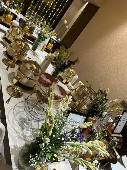 A long shot of an elegant buffet line ready for a house party. The setup includes our signature gold serving dishes, beautiful floral arrangements, and ambient lighting to create a sophisticated dining experience at home.