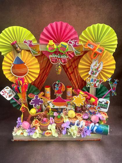 A grand Holi hamper designed with a colorful backdrop of paper fans. This arrangement is overflowing with festive goodies, from pichkaris and colors to sweets and snacks, making it the ultimate Holi gift.