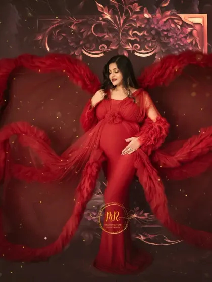 A dramatic solo portrait in a deep red gown with feathered details. The fabric is styled to create a wing-like effect against a rich, textured backdrop, perfect for a bold pregnancy announcement.