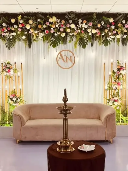 A refined and elegant stage setup featuring a lush floral header, warm candle-lit stands, and a traditional brass lamp (samai) in the foreground. This design beautifully merges modern aesthetics with timeless traditions.