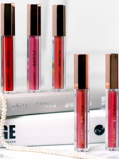 A clean, minimalist arrangement of liquid lipsticks resting on a stack of books. The composition is simple yet elegant, using a string of pearls to add a classic touch to the modern product design.