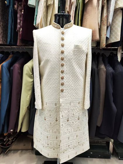 An elegant white sherwani on a mannequin, featuring a textured fabric and an embroidered collar. The gold buttons add a touch of regality to the classic design.