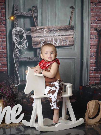Howdy, partner! This little cowboy is all smiles on his rocking horse at the "Father's Ranch." The rustic brick and wood background, complete with a cowboy hat and lasso, sets the scene for a perfect Wild West themed shoot.