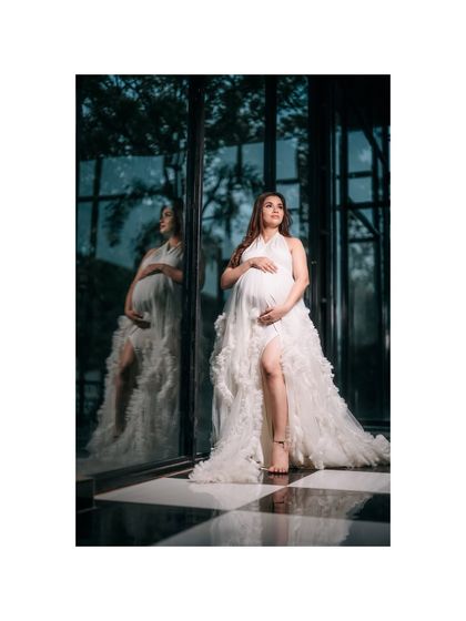 A stunning full-length portrait of a mom-to-be in a white gown, standing against a reflective glass wall. Her reflection adds a beautiful, artistic element to this outdoor maternity shot.