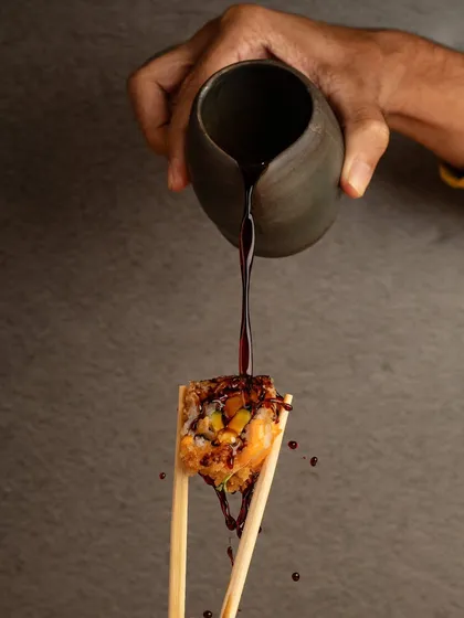 A dramatic action shot of soy sauce being poured over a piece of sushi held by chopsticks. The splash and motion create a dynamic and high-end feel.