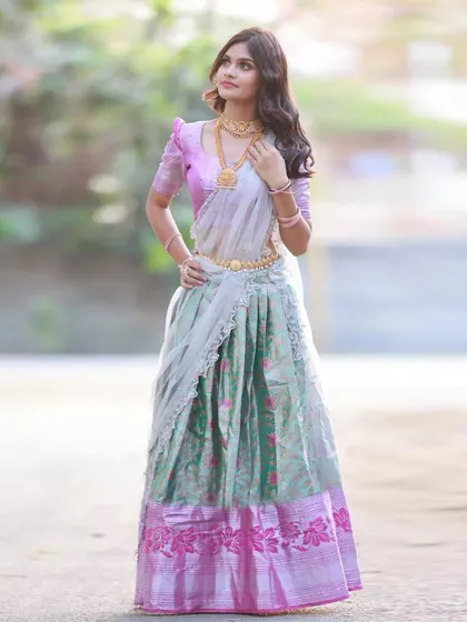 A beautiful half-saree in pastel shades of mint green and pink. This full-length outdoor shot showcases the entire outfit, including the intricate border and the elegant drape of the dupatta.