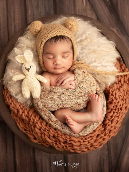 A cozy, rustic theme using warm, earthy tones. The baby, wearing an adorable bear bonnet, is curled up in a bowl with a knitted wrap and a small rabbit friend.