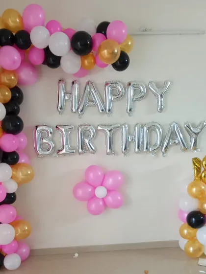 A classic and simple home birthday decoration. This L-shaped balloon arch in pink, black, white, and gold is paired with silver 'Happy Birthday' foil letters and a number balloon column, perfect for an 18th birthday party at home.