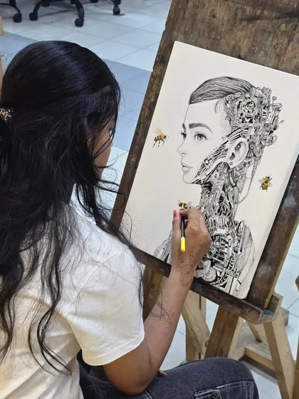 A student works on her surreal portrait, using a micron pen to draw complex mechanical details emerging from the subject's neck and head.