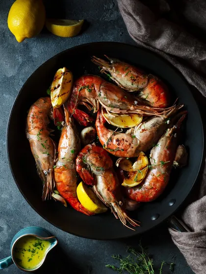 A dark and moody shot of butter garlic prawns for The Khyber Resort. The overhead lighting creates dramatic shadows, emphasizing the rustic pan and the vibrant pink of the cooked prawns with lemon.