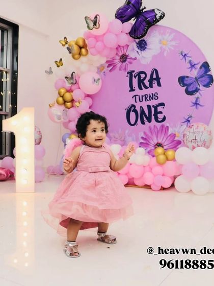 A precious moment captured at a butterfly-themed first birthday party. The birthday girl stands before a purple circular backdrop decorated with butterflies and flowers, with a large light-up number '1' beside her.