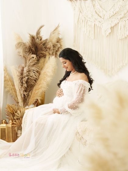 A dreamy portrait of an expecting mother in a white, pearl-studded gown. She is seated in a soft, light-filled boho setting, looking down at her bump with a peaceful smile.