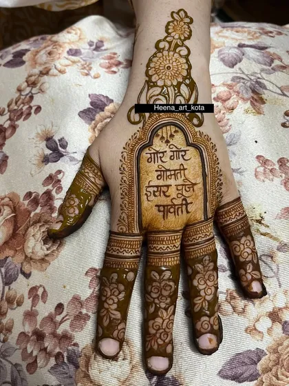 A simple and traditional Gangaur mehndi. The palm features the sacred mantra "Gor Gor Gomti" inside an ornate frame, with floral details on the fingers.