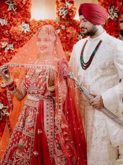 A classic portrait of a Sikh couple on their wedding day. The groom, holding the kirpan, stands proudly with his bride, who lifts her veil with a smile, against a stunning red floral backdrop.