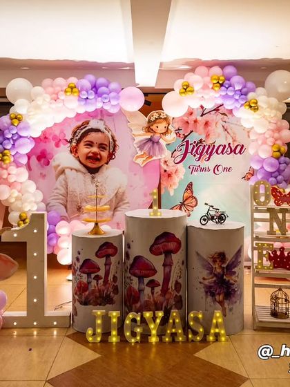 A fairy and butterfly-themed first birthday with a personalized backdrop featuring the child's photo. The decor includes themed plinths with mushroom and fairy graphics, a light-up number '1', and a pink and purple balloon arch.