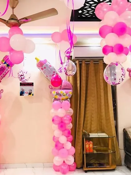 An elaborate pink and white balloon decoration for a baby girl's welcome home party. The setup includes two large balloon pillars topped with baby bottle foil balloons and clusters of balloons on the ceiling.