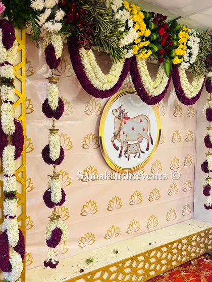 A beautiful backdrop setup featuring the sacred cow motif, framed by a golden jaali and hanging floral arrangements. The pink background with lotus prints adds a delicate touch.