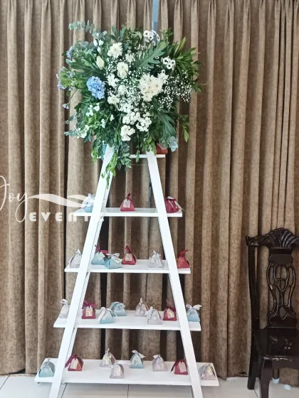 A creative display stand for return gifts at a baptism ceremony. We used a white ladder shelf, topped with a large floral arrangement, to present the party favors in an organized and attractive way.