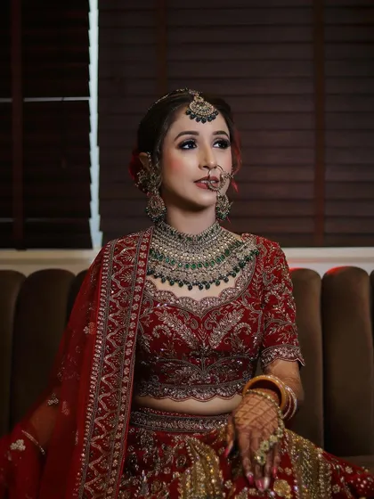 A full-length view of the bride seated, showcasing her complete look. The makeup is perfectly balanced with her elaborate red lehenga and heavy jewelry, creating a regal and timeless bridal appearance.
