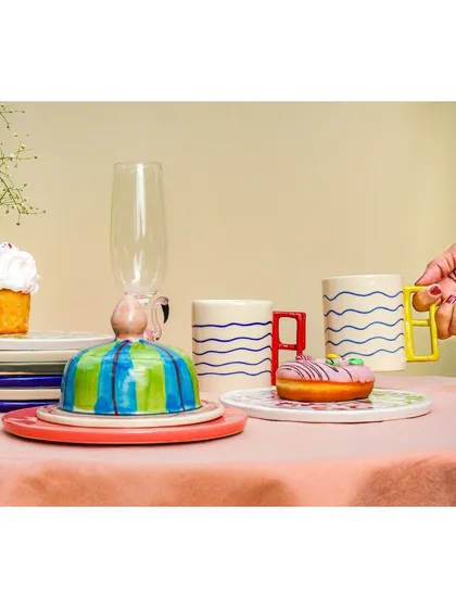 A lifestyle product shot with a hand reaching for a quirky, modern mug, part of a colorful tableware collection.