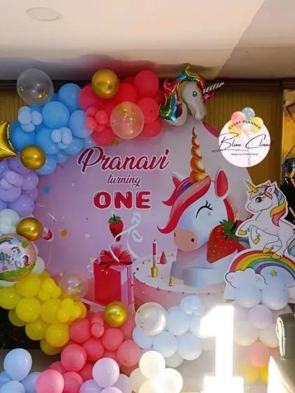 A magical unicorn-themed first birthday party. The decor features a custom backdrop with unicorns and rainbows, surrounded by a colorful pastel balloon garland.