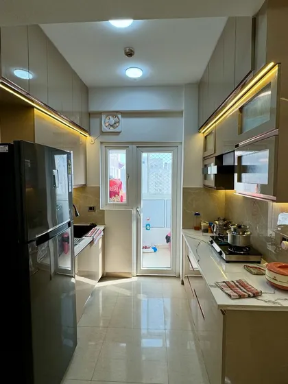 A look inside a completed 2BHK apartment kitchen in Noida Extension. The glossy beige cabinets are complemented by rose-gold profile handles and fluted glass shutters.