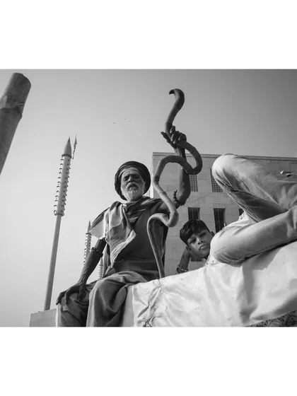 A low-angle black and white shot of a sadhu in Varanasi, holding a staff with a sculpted snake. A young boy peeks out from behind him, adding a layer of curiosity to the scene.