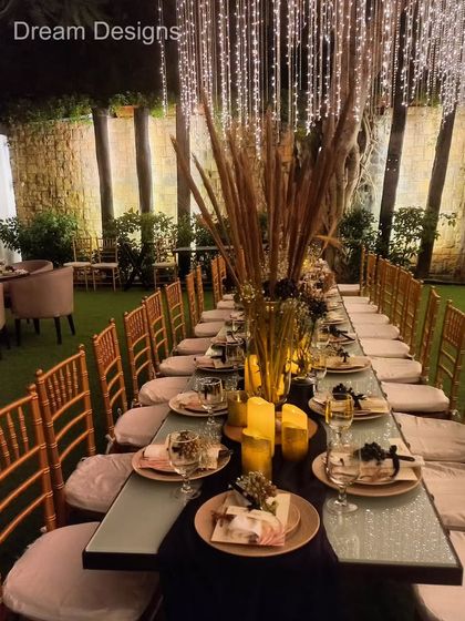 A formal sit-down dinner with a rustic touch. The long table is decorated with tall vases of pampas grass and warm candles, set under a canopy of hanging lights for a magical "dinner under the stars" feel.