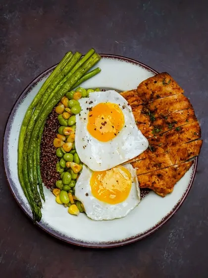 A healthy and high-protein meal with grilled chicken breast, sunny-side-up eggs, quinoa, and green beans. The dark, dramatic lighting and top-down view create a powerful and appetizing fitness meal shot.
