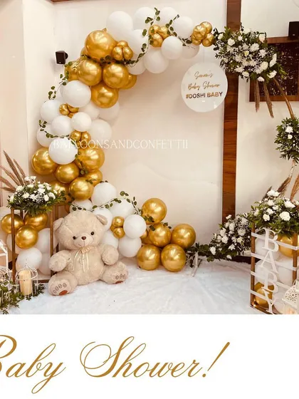 A classic and elegant baby shower theme with a circular arch of white and gold balloons. The teddy bear and floral arrangements add a soft, sweet touch.