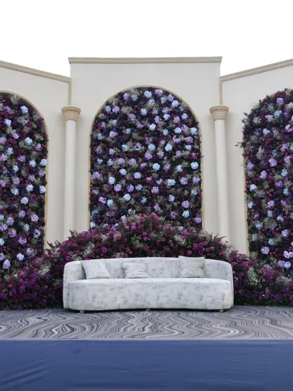 A beautiful stage backdrop featuring three arches filled with a dense arrangement of purple and blue flowers, creating a lush and elegant photo wall.