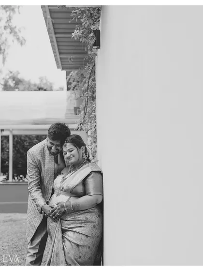 A tender black and white photograph of an expecting couple in a quiet embrace against a clean white wall. The simplicity of the composition draws all attention to their connection.
