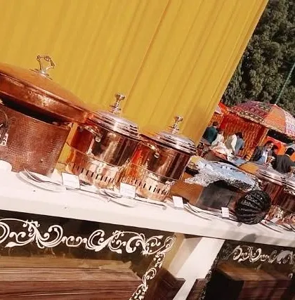 A vibrant buffet setup for a daytime event with a bright yellow backdrop. The table features traditional copper serveware and intricate white decorative elements on the counter front.