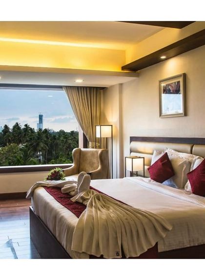 A hotel bedroom in Calicut designed for comfort and luxury. The room features a large window with expansive views, soft ambient lighting from the ceiling cove, and a well-appointed bed.