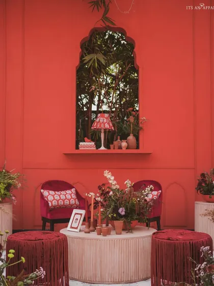 An intimate seating nook within the 'Gulal Bagh' theme, featuring a rich red backdrop, velvet armchairs, and custom-printed textiles.