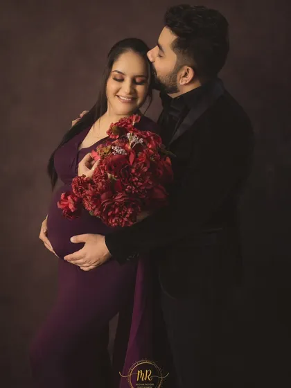 A romantic couple's portrait with a floral touch. The partner embraces his wife, who is holding a bouquet of red flowers that matches her deep purple gown.