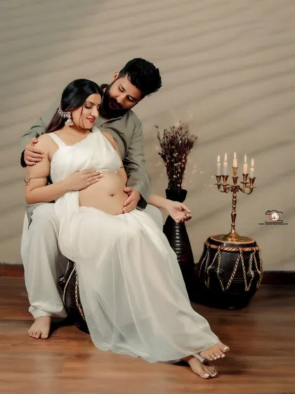 A tender moment between partners during a traditional-themed maternity shoot. The expecting mother, dressed in a simple white saree, is seated on her partner's lap as they both look down at her baby bump.