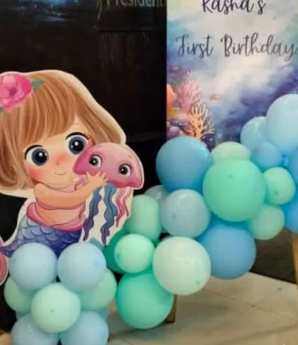 A cute mermaid character cutout placed next to a personalized welcome board for a first birthday party, greeting guests as they arrive.