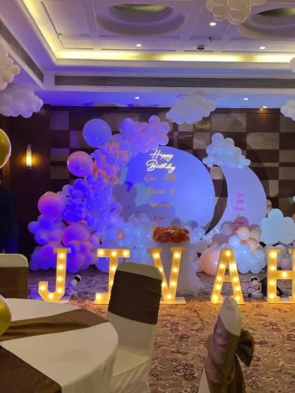 A grand birthday celebration in a banquet hall with large, illuminated marquee letters spelling out the name 'JIVAH'. The backdrop is a dreamy moon and clouds theme, created with custom panels and extensive balloon work.