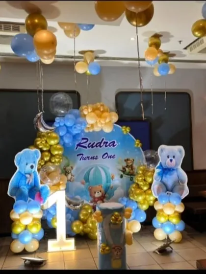 A sweet and cuddly teddy bear theme for a first birthday. This decoration features a "Rudra Turns One" backdrop, large bear cutouts, a light-up number '1', and a blue and gold balloon arch.